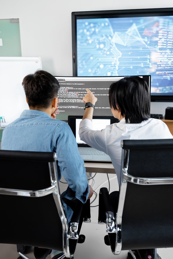 Home Two adults collaborating in an office, discussing code on a computer screen.